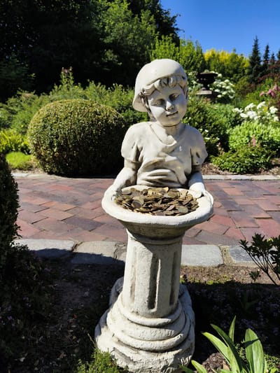 This media file shows a statue of a young child wearing a cap, holding a bowl filled with coins. The statue is made of a light-colored material, possibly stone or concrete, and stands on a decorative pedestal. The child appears to be looking down at the coins with a slight smile.
The statue is situated in a garden setting. In the background, there are various types of greenery, including a large, rounded bush, trees, and flowering plants with white and pink blossoms. A brick pathway runs behind the statue. The sun is shining brightly, casting shadows, suggesting it is daytime. The clear blue sky indicates fair weather.
The overall scene evokes a sense of playful generosity or perhaps a wishing well. The presence of the coins suggests people have been interacting with the statue, possibly making wishes or donations. The garden environment implies an outdoor public space, such as a park, botanical garden, or the grounds of a historical site. The provided location context places this scene in Goczałkowice-Zdrój, Poland. There is no visible text in the image.