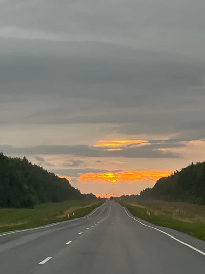 This is a vertically oriented, eye-level shot of a paved road leading into the distance. The road has white dashed lines marking its lanes and is bordered by a grassy shoulder and dense green trees on the left and a less dense line of trees and grass on the right.
The sky is a dramatic canvas of clouds. On the left, the clouds are a uniform, heavy gray, suggesting overcast or impending rain. However, on the right, the clouds are illuminated by a vibrant sunset, displaying fiery oranges and yellows. This contrast between the dark, stormy clouds and the brilliant sunset creates a striking visual.
The time of day is clearly sunset, evidenced by the colors in the sky. The weather appears mixed, with a potential for rain on the left and clear, vibrant sunset skies on the right. The scene evokes a sense of journey or travel, with the road stretching towards the horizon where the sky is ablaze. The presence of trees suggests a rural or semi-rural location. No people or text are visible in the image.