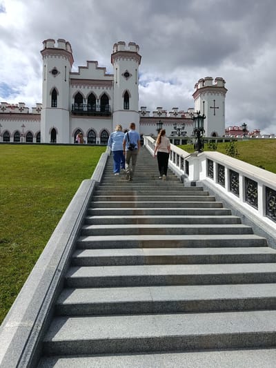 The image shows a wide, granite staircase leading up to a white, castle-like building with pink accents, situated in Kosava, Belarus. Three individuals are ascending the stairs. In the foreground, a woman with blonde hair, wearing a blue shirt and dark pants, is in the lead. Behind her is a man in a light blue shirt and khaki pants, carrying a black satchel. Further up the stairs and slightly ahead is a woman with long, brown hair, wearing a beige shirt and black pants. The building has multiple turrets and arched windows, with a large expanse of green lawn to the left of the staircase. The sky is cloudy with patches of blue.