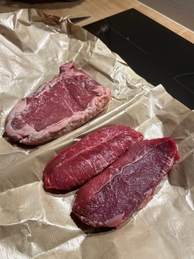 The image displays three raw cuts of meat resting on a crumpled sheet of brown butcher paper, positioned on a light-colored wooden countertop. In the upper left quadrant, a single large, raw, bone-in steak, consistent with a T-bone or similar cut, exhibits red muscle tissue, visible marbling, and a thick, pale fat cap along its perimeter. Towards the lower right, two smaller, boneless cuts of raw red meat are present, appearing leaner than the larger steak. In the background, a dark, reflective induction cooktop is partially visible on the countertop, along with the tip of a knife blade. No individuals are present. The precise location within the Archanes Municipal Unit, Greece, cannot be determined from the visual information provided.