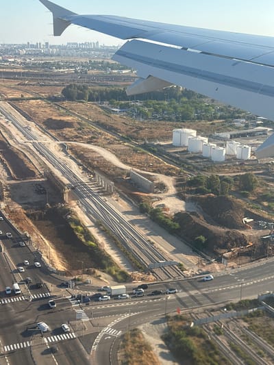 This is an aerial photograph taken from an airplane window, looking down at a landscape that appears to be undergoing development or construction. The perspective is tilted, with the wing of the airplane occupying the left side of the frame. The scene below reveals a mix of industrial structures, natural terrain, and infrastructure.
Prominent subjects include several large, white cylindrical tanks, possibly for storage, arranged in a line. Adjacent to these, there are extensive railway tracks and what appears to be a construction site with earthmoving equipment and piles of dirt. A road with several cars is visible, indicating ongoing traffic and activity. In the background, a cityscape with numerous buildings suggests a populated area.
The setting is likely a semi-urban or industrial zone bordering a more developed city. The landscape is dry and somewhat arid, with patches of green vegetation interspersed with brown, uncultivated land. The time of day appears to be daytime, with clear skies and bright sunlight casting distinct shadows, suggesting fair weather.
There is no visible text in the image. The overall impression is one of active development and infrastructure projects in a warm, sunny climate. The presence of the airplane wing signifies travel and a high vantage point, offering a broad overview of the area.