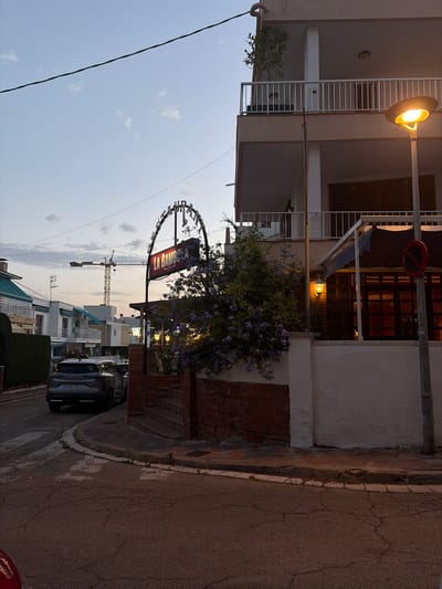 In the city of Cambrils Badia, Spain, at dusk, a street corner is captured. A silver SUV, license plate 2491 NCT, is parked on the left side of the road. Adjacent to the road, a brick wall encloses a property, topped with white plaster. A bush with small purple flowers blooms beside the wall. A brick staircase leads up to a building with dark-paneled windows and a dark awning. Above the entrance, a sign arching over the walkway reads "LA BAMBOLA RESTAURANT." A street lamp, illuminated with a warm yellow light, stands near the sign. A no-parking sign is visible on a pole. In the background, a construction crane rises against the twilight sky, with residential buildings and a power line also present. The street is paved with asphalt showing signs of wear and cracking.