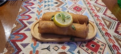 Three golden-brown, crispy rolls are presented on a small, round wooden plate. A slice of lemon, garnished with green herbs, sits atop the rolls. The presentation is set against a patterned cloth with geometric shapes in brown, blue, orange, and red hues. The backdrop includes a brown surface, possibly a wooden table. A fork is partially visible in the upper left corner.
