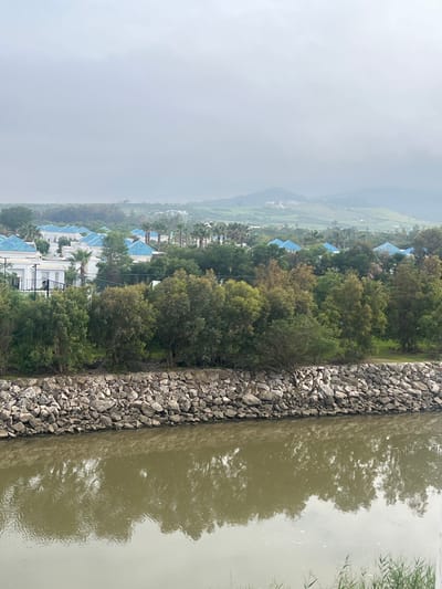 The image shows a view of Fnideq, Morocco on a hazy day. The scene includes a body of water, possibly a river, with a rocky embankment on one side. The water is reflecting the sky and trees. Adjacent to the riverbank are trees and a row of buildings with blue tiled roofs. The landscape further includes rolling hills visible in the background, shrouded in mist or low clouds. The general atmosphere is overcast and damp.