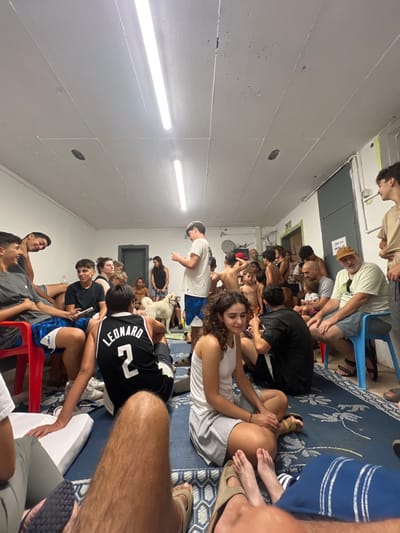 A crowded room features a diverse group of people, some sitting on chairs and others on a patterned carpet. Fluorescent lights illuminate the space, exposing white walls. Several individuals are engaged in conversations or activities, while others are focused on personal devices. A few people are shirtless. A dog is sitting among the seated people. The scene suggests a casual, informal gathering.