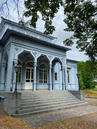 The image depicts the exterior of a light blue wooden building, likely a historic house or museum, in Sortavala, Russia. The building features intricate architectural details, including decorative trim, arched verandas with columns, and large windows. A wide, stone staircase with multiple steps leads up to the main entrance, which is marked by a white double door. Two black lampposts stand on either side of the staircase. The building is surrounded by greenery, with trees and foliage visible in the foreground and background. The sky is overcast, suggesting a cloudy day. The overall impression is one of old-world charm and historical significance.