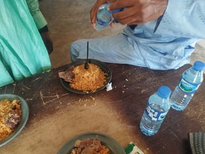 The image shows a wooden table with three plates of food, three bottles of water, and a person's hand holding a bottle of water. The person is sitting on a chair, wearing a long blue shirt and a pair of pants. They are pouring water from the bottle into their hand. Two of the water bottles are "Sweet" brand and the other is unlabeled. One plate of food contains rice and meat, and the other two plates contain rice and meat. The image appears to be taken in Kaltungo, Nigeria, based on the provided coordinates (latitude: 9.816, longitude: 11.315). The scene appears to be a casual meal in a private or semi-private setting.