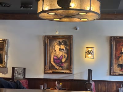 The image depicts the interior of a restaurant, likely a bar or steakhouse, with a rustic and elegant ambiance. The foreground shows a table with several glasses and a bottle of ketchup, suggesting a dining setting. In the mid-ground, a large framed painting of a couple, a man in a cowboy hat and a woman in a purple dress, is prominently displayed on the wall. To the right of this painting, a wall sconce with a "W" logo emits a warm light. The background features more framed artwork and dark wooden paneling on the walls, consistent with a Western-themed establishment. The ceiling has a large, ornate light fixture. The overall scene suggests a well-decorated and possibly upscale dining or entertainment venue. The location is identified as Jackson, United States of America, though the specific establishment within Jackson is not detailed in the visual information.