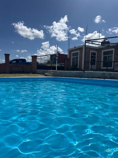 The image depicts a bright, sunny day in San Baltazar Torija, Mexico, featuring a swimming pool in the foreground. The pool's water is a vibrant blue, with sunlight creating shimmering patterns on its surface. In the background, a red brick building with white-trimmed windows and doors is visible. A black metal fence separates the pool area from the building and yard. A blue pickup truck is parked behind a brick wall and a black metal fence to the left of the frame. The sky is a clear blue with scattered white clouds. The scene appears to be a residential or recreational property.