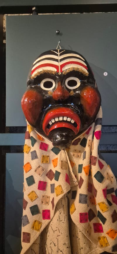 The media file shows a close-up, overhead view of a traditional black puppet mask with a wide, red, toothy smile and white eyes, displayed against a light-colored, textured fabric adorned with colorful diamond and square patches. The mask is the primary subject, with its glossy black surface contrasting with the vibrant red of the mouth and cheeks, and the stark white of the eyes and facial markings. The fabric appears to be draped around the mask, suggesting it might be part of a costume or display.
The setting is indoors, likely a museum or exhibition space in Sargans, Switzerland, given the context. The background is a muted, dark gray, possibly a wall or display case, with a hint of a dark metal structure at the bottom of the frame, indicating the object is on display. There is a small, white circular emblem visible on the gray background to the left of the mask, possibly a museum label or identifier, though no visible text is legible. The lighting is even, highlighting the textures and colors of the mask and fabric without harsh shadows. The overall impression is a still, curated display of a cultural artifact.