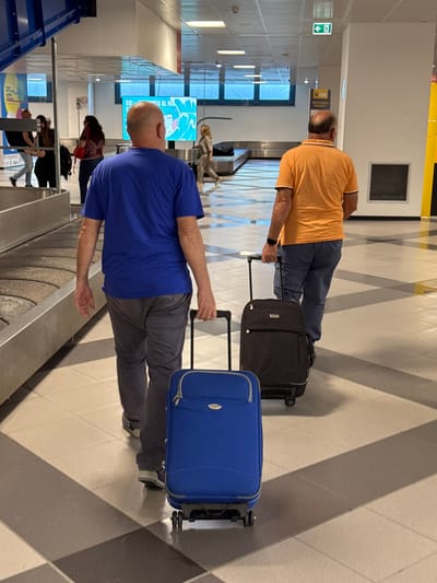 The image depicts two men at the baggage claim in an airport in Cinisi, Italy. One man wears an orange shirt and dark pants, pulling a black suitcase. The other man wears a blue shirt and gray pants, pulling a blue suitcase. They are positioned near a baggage carousel. Other people are visible in the background. The floor has a geometric pattern. Signage is visible on the walls, and a blue structure is present on the left side of the image.