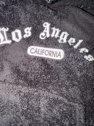 The image shows a close-up of a black, fuzzy fabric with white lettering that reads "Los Angeles" and "California." The lettering appears to be stitched onto the fabric. The background is out of focus, but appears to be more of the same fuzzy material. There is no indication of location in the city of Ivanski, Bulgaria, in the image.