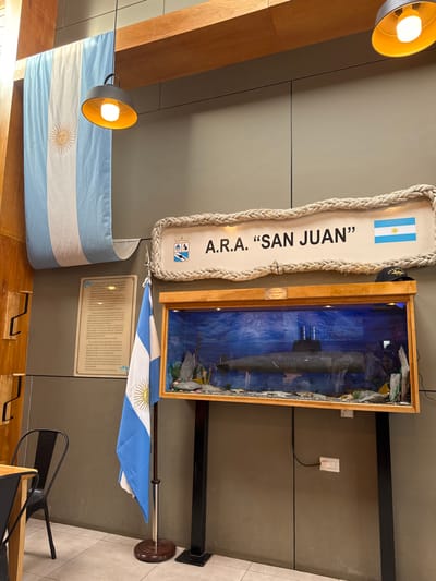 The scene depicts an interior space, identified as being in the city of Tolhuin, Argentina, featuring a memorial display dedicated to the A.R.A. San Juan submarine. The background consists of plain light gray walls and a tiled floor. Overhead, light-colored wooden beams traverse the space, from which two black, inverted bell-shaped pendant lights hang, each emitting warm light.
Prominently displayed on the main wall is a large, horizontally oriented sign with a thick rope border. The sign's white face reads "A.R.A. 'SAN JUAN'" in dark, capitalized letters, flanked by a crest on the left and a small Argentine flag on the right. Below this sign, a rectangular wooden-framed display case, approximately 1.5 meters wide, showcases a detailed model of a submarine, likely the A.R.A. San Juan, against a blue background mimicking water, with artificial rock formations and marine plants at its base. This display case rests on two vertical dark metal posts. On top of the display case, a dark cap with "A.R.A. SAN JUAN" embroidered on it is visible towards the right side.
To the left of the submarine display, an Argentine flag is presented on a standing pole with a circular wooden base. Above and to the left of this flag, a rectangular informational plaque with text is affixed to the wall. Further left, a wooden panel with multiple hooks (a coat rack) is mounted. A large Argentine flag is draped vertically from the upper wooden beam on the far left, descending towards the floor. In the bottom left corner, part of a wooden table and a black metal chair are visible. A faint reflection of a person is discernible in the glass of the submarine display case. A white electrical outlet and a black cable are visible on the wall behind the right support post of the submarine display. No active people or specific interactions are observed.