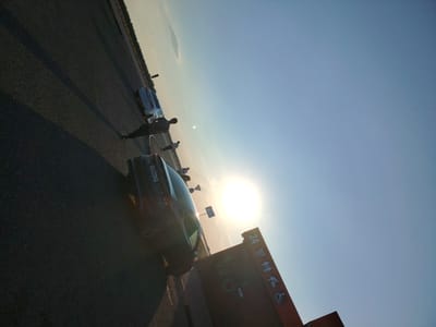 The image shows a street scene in the city of Arshaly, Kazakhstan, during sunset. A dark-colored car is parked in the foreground on the left, with its license plate visible. In the middle ground, several people are standing near two white cars. The sun is setting on the right side of the image, casting a bright glare. A large, red sign with white symbols and text is also visible on the right, possibly indicating a service area or parking lot. The ground appears to be asphalt, and there are shadows cast by the cars and people. The sky is clear and blue, transitioning to a lighter hue near the horizon.