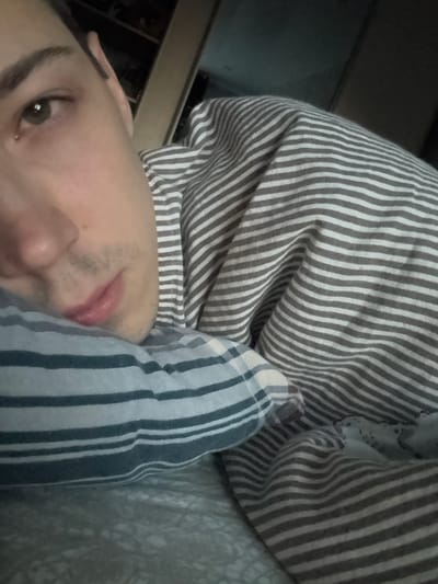 A close-up, overhead shot shows a person lying in bed, facing left. Their head rests on a light blue and white striped pillow. Their face is partially visible, with their left eye, nose, and mouth in focus. They are wearing a grey and white striped shirt or pajamas, which are draped over them. The background is dark and out of focus, with a hint of shelves and objects on the right. The image is indoors, likely in a bedroom. The location is Alexeyevskoye, Russia.