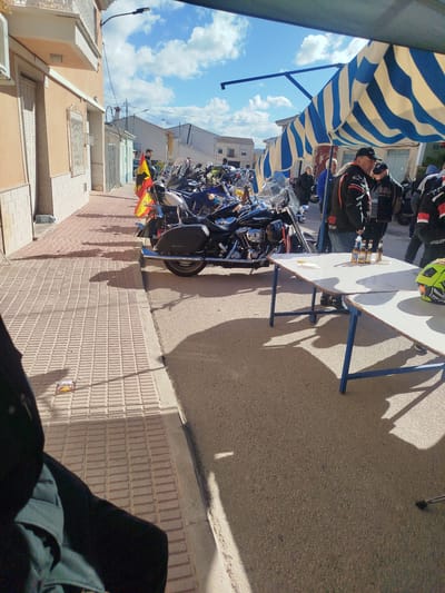 A scene in Mula, Spain, shows a street lined with parked motorcycles. A table with bottles and a helmet sits in the foreground. Several people stand around the table and motorcycles, suggesting a gathering. Spanish flags are visible on some of the motorcycles. The environment includes buildings lining the street, a striped awning, and a clear sky. The lighting indicates a sunny day.