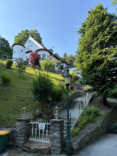 The image shows a white house with rounded towers on a grassy hillside in Bad Gastein, Austria. A stone staircase with a white metal railing leads up the hill towards the house. A vintage green street lamp stands next to the staircase. There are trees and bushes surrounding the property. A white gate with a "Privat" (private) sign is visible at the bottom of the stairs. The sky is clear blue with a few wispy clouds.