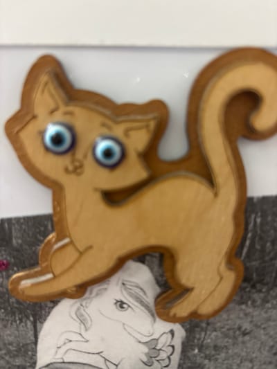 The image shows a close-up of a wooden cutout of a cat. The cat is a light brown color and has a stylized design. It appears to be in a playful pose, with its tail curved upwards and its body stretched out. The cat has two round, blue eyes that resemble evil eye charms. A faint, cartoonish smile is visible on its face. Behind the cat cutout, there is a drawing of a face, possibly an elephant or a mythical creature, rendered in black and white pencil. The background is a mix of blurry textures, with a gray, textured surface on the right and a plain white surface on the left. The lighting is bright, suggesting it was taken indoors. No specific time of day, weather, or explicit location cues are discernible beyond what might be inferred from the general setting of a home or craft space. No visible text is present.