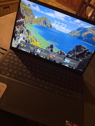 A black Dell Inspiron 15 laptop, identifiable by its model label and AMD Ryzen/Radeon stickers on the palm rest, is depicted from an oblique, elevated perspective. The laptop rests on a textured, light brown surface, likely a table. Its screen is active, displaying the Windows lock screen with a background image of a tropical lagoon surrounded by rocky, vegetated islands. The digital clock on the screen reads 22:22, and the date is "miércoles, 25 de febrero." A weather widget shows "Zanouverde, 11°C," described as "Prácticamente despejado," with temperature ranges of 11°/19°. Below this, several news widgets in Spanish are visible, including "La maravilla de cada día," "Este día en la historia," and "Cuestionario diario." A Wi-Fi signal icon and a battery indicator showing 32% charge are present in the lower right corner of the screen. The laptop's black keyboard features a QWERTY layout with a numeric keypad, and a large central touchpad is visible. The scene is illuminated by dim, ambient light, suggesting an evening setting, with blurred, dark elements in the background. The described location is Acassuso, Argentina.