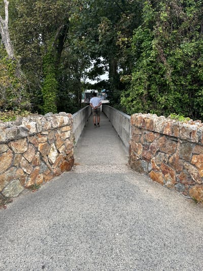 A person is walking away from the camera on a paved pathway that is flanked by stone walls on either side. The pathway leads towards a bridge with a metal railing. The stone walls are constructed from irregularly shaped, reddish-brown stones. The pathway is surrounded by dense green foliage and trees, suggesting a natural or park-like setting. The person is wearing a white t-shirt and khaki shorts, with their hands clasped behind their back. In the distance, a white van and a bicycle can be seen. The scene appears to be in Courtown, Ireland, possibly near a coastal area given the natural elements.