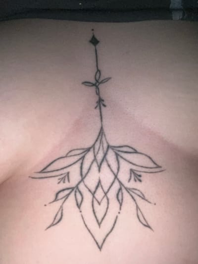 The image shows a close-up of a black linework tattoo on a person's chest. The tattoo is symmetrical and extends vertically, with a diamond shape at the top and a floral or botanical design at the bottom. The floral design features flowing lines and leaf-like elements. The skin appears to be fair. The background is dark and out of focus, suggesting the image was taken indoors. There is no discernible indication of the city of Markova, Russia, or any other environmental details beyond the immediate subject.