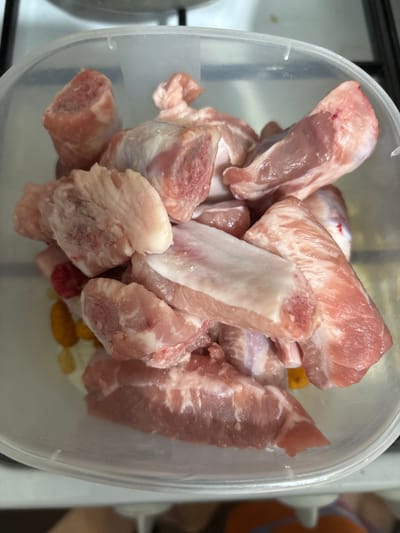A close-up, overhead shot shows a clear plastic container filled with raw pork ribs. The ribs are cut into smaller pieces, some with bone and others with more meat. The meat is a light pinkish-red color, with visible layers of fat and muscle. Some pieces show evidence of a rough cut, with jagged edges. At the bottom of the container, a few small, yellowish objects are visible, possibly spices or seasonings. The overall lighting is bright, suggesting an indoor setting, likely a kitchen, given the glimpses of white stovetop elements and a tiled surface in the background. The scene is static, with no activity depicted beyond the preparation or storage of the raw meat. There is no visible text in the image.