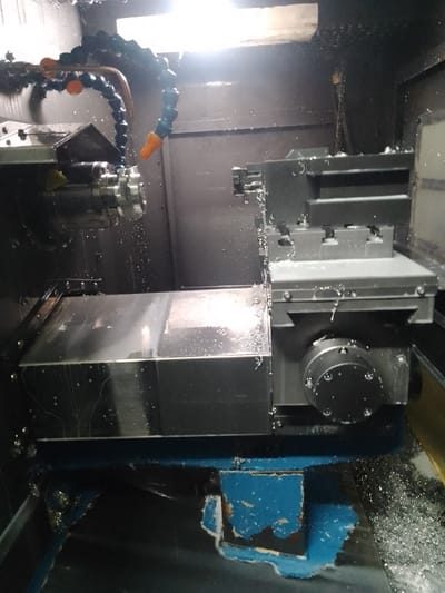 This media file shows a close-up view of a metal-cutting machine, likely a CNC lathe, in operation. The scene is inside a workshop, suggested by the industrial machinery and the presence of metal shavings. A coolant hose is visible, spraying a liquid onto the workpiece and cutting tool to keep them cool during the machining process. The machine itself appears to be made of dark metal and features various components such as a chuck, a cutting tool holder, and a movable carriage. Metal shavings are scattered around the base of the machine, indicating that material is actively being removed from a workpiece. The lighting suggests an indoor, possibly artificial, light source, and the overall impression is one of active manufacturing. There is no visible text in the image. The location context provided is Ciantra, Indonesia, which places this industrial activity in a specific geographical setting.