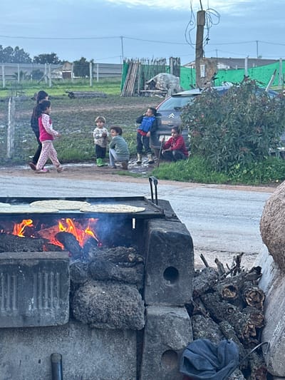 A group of children stand near a car on a dirt road adjacent to a grassy field, while two individuals watch nearby. In the foreground, a fire burns inside a brick stove, with tortillas cooking on top. Wood and a blue jacket are stacked behind the stove. A utility pole stands in the distance, and a gray, cloudy sky is overhead.