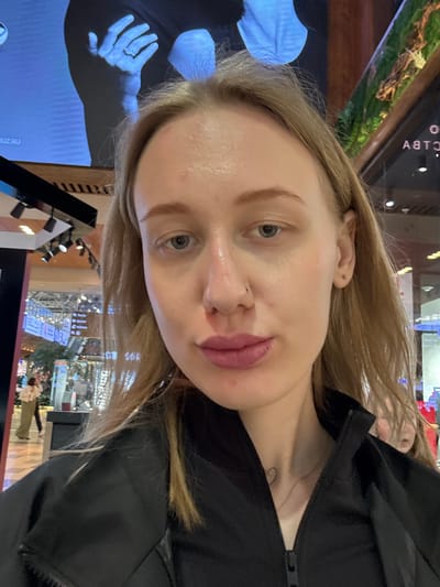 A selfie of a fair-skinned woman with blonde hair, blue eyes, and a nose piercing. She is wearing a black jacket. A tattoo is visible on her chest. The background appears to be a public space with a display screen showing an image of a hand. Other displays and interior architecture are visible. The location is not identifiable as Ol'ginskaya, Russia based on the provided information.