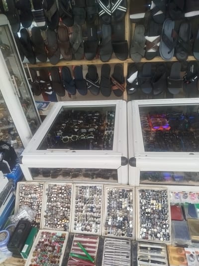 The image depicts a street market stall in Way, Nigeria, displaying a variety of accessories. The stall is organized with shelves and display cases. On the upper shelves, numerous pairs of sandals and flip-flops are arranged in rows. Below, large glass display cases showcase a collection of sunglasses and eyeglasses. Some of the eyeglasses have tinted lenses. Further down, several shallow boxes contain an assortment of small items. These include what appear to be cuff links or tie clips with geometric patterns and various button-like accessories, some with intricate designs. Other boxes contain what seem to be rings or brooches with colorful stones. The bottom of the display features pens, including fountain pens and rollerball pens, as well as what might be small electronic devices or charging accessories. The overall impression is one of a busy market stall selling a wide range of personal accessories and small items. The lighting suggests it is daytime, and the setting appears to be an open-air or semi-covered market.