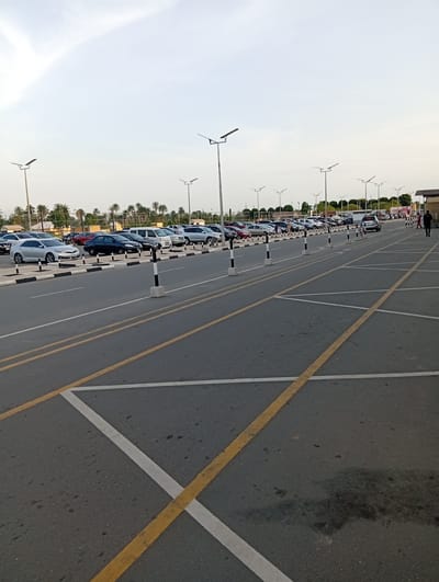 The image shows a large, mostly empty parking lot in Omagwa, Nigeria. Rows of parked cars are visible in the background, separated from the empty parking spaces by a row of bollards. Overhead are tall lampposts. The parking spaces are demarcated with yellow and white lines on the asphalt. A person is walking in the background near a building. The sky is overcast.