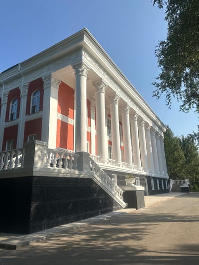 The image shows a grand building with a white colonnade and red walls. The building has classical architectural elements, including decorative moldings and ornate capitals on the columns. There is a wide staircase leading up to the entrance, adorned with a white balustrade. The base of the building is made of dark, rough-textured stone. In the foreground, a paved pathway runs alongside the building, with shadows from trees cast upon it. The sky is clear and blue, and mature trees are visible in the background, suggesting a park-like setting. The overall impression is one of historical significance and architectural beauty. This building is the sanatorium "Ust-Kachka" in the village of Ust-Kachka, Perm Krai, Russia.