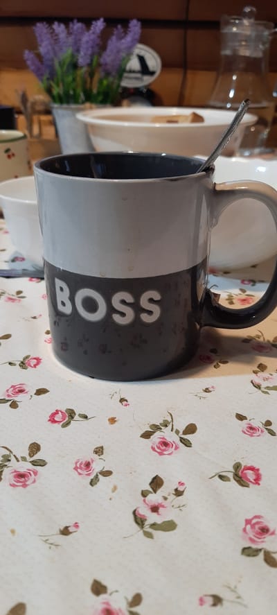 A close-up shot focuses on a dark gray mug with the word "BOSS" emblazoned in white letters. The mug is two-toned, with the upper portion being a lighter gray. A silver spoon is partially visible inside the mug, resting against the rim. The mug sits on a white tablecloth adorned with a pattern of small pink roses and green leaves. In the blurred background, a glass pitcher filled with liquid can be seen, along with a white bowl, and a vase with purple flowers. The setting appears to be indoors, possibly a home or cafe, with wooden elements visible in the background.