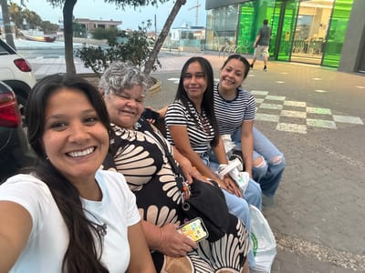 In Garrucha, Spain, a group of four individuals, likely a family, are seated on a bench outside a building with green glass windows. The woman in the foreground, on the left, is taking a selfie, smiling warmly at the camera. She is wearing a white t-shirt. To her right is an older woman with gray hair, also smiling and looking towards the camera. She is wearing a black and white patterned dress. Next to her are two younger women, both wearing striped t-shirts and ripped jeans. The younger woman on the left is looking at the camera and smiling, while the woman on the right is also smiling and looking at the camera. In the background, a man in a dark shirt and shorts is walking away from the camera towards the building. The scene appears to be in a public area, possibly a promenade or street, with a sidewalk and a street in the background. There is a car visible on the left side of the image and a building with large green windows on the right. The lighting suggests it is late afternoon or early evening.