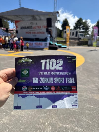 This media file shows a close-up of a hand holding a race information pamphlet for the "16K-ZORKUN SHORT TRAIL" race, likely part of the "AMANOS ULTRA TRAIL" event. The pamphlet features the number "1102" and the name "VITALII GONCHARUK." It includes a course profile with elevation markers and cut-off times for checkpoints and the finish line. Visible text on the pamphlet also includes sponsor logos like "GARMIN," "ideal," "KARTAL," and "BADE GAZOZ." Additionally, it lists emergency contact numbers and the location "OSMANİYE -SPOR KULOBO-".
In the background, a blurry scene suggests an outdoor event with a stage or tent structure, potentially a starting or finishing area. People are visible in the background, some gathered around the stage, while others are more dispersed. The overall setting appears to be a race event, possibly in Turkey given the text and location cues. The lighting suggests it is daytime, with clear skies and sunlight casting shadows, indicating fair weather. The focus on the pamphlet implies the viewer is likely a participant or spectator interested in the race details. The overall tone is informative and pertains to an athletic competition.