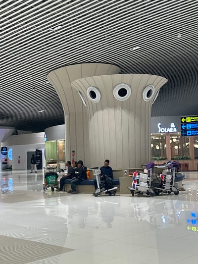 The image depicts the interior of an airport terminal in Baji Mangngai, Indonesia. A group of people are seated on a bench in the foreground, waiting with their luggage. There are several luggage carts loaded with bags. To the left, a person is walking away from the camera. The architecture features a unique, large, light-colored structure with circular openings, resembling eyes, overhead. The ceiling is composed of parallel black and white stripes. Signage is visible in the background, indicating directions to "Karang", "Hotel", and "Mushola". The floor is highly reflective, showing distortions of the surroundings. The overall scene suggests a typical airport waiting area.