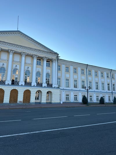 A low-angle, full shot captures the exterior of a historical building in Yazvitsevo, Russia, under a clear blue sky. The building is a grand, multi-story structure with a pale yellow facade on its upper levels and white on the lower sections. The left side of the building features a prominent portico with large, white Ionic columns supporting a triangular pediment. White lettering in Russian is visible on the pediment. Numerous tall, arched windows with black railings are present on the upper floors. The ground level of this section has two large, wooden double doors.
The building extends to the right with a longer, more uniform facade featuring rows of rectangular windows with white frames. A double-headed lamppost stands in front of this section, flanked by small evergreen trees. The street in front of the building is paved with asphalt and marked with white lane lines. The scene appears to be a quiet, urban environment. No people are visible in the image. The time of day is likely late afternoon or early morning, given the quality of the light and the clear sky.