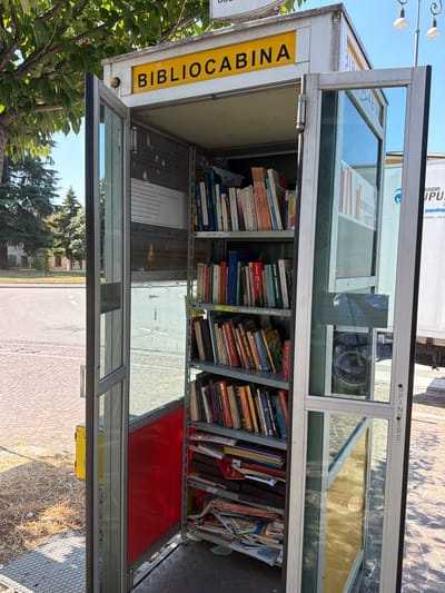 The image depicts an outdoor "bibliocabina" in Udine, Italy. This is a public book exchange, housed in a structure resembling a repurposed telephone booth. The structure has glass doors and sides, revealing shelves filled with books. The "BIBLIOCABINA" sign is prominently displayed in yellow lettering against a white background on the top of the structure. The surrounding environment shows a paved area with some dry grass and trees. In the background, a white truck is partially visible, suggesting an urban or semi-urban setting. The weather appears to be sunny with clear blue skies.