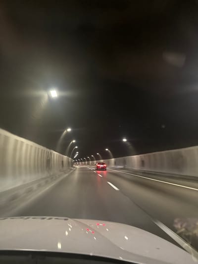 The image depicts a view from inside a vehicle traveling through a tunnel. The tunnel is illuminated by overhead lights, and its walls are lined with what appear to be concrete barriers. Multiple cars are visible ahead, their taillights glowing red. The road surface is wet, suggesting recent rain or humidity. The foreground shows the hood of the vehicle from which the photo is taken. The scene is consistent with a highway tunnel, and the prompt specifies the location as Dolna Vasilitsa, Bulgaria.