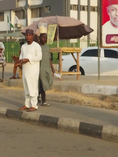 This image captures a daytime street scene in Gyadi Gyadi Arewa, Nigeria, with a soft, ambient light suggesting late afternoon. In the foreground, a man stands centrally, facing forward, dressed in a white traditional long-sleeved tunic (likely a kaftan or similar attire) and trousers, complemented by a dark cap (kufi). He wears brown sandals and appears to be holding a small item in his hands.
Behind him and slightly to his right, another individual is partially visible, possibly walking, with a white, circular container balanced on their head, a common method of carrying goods in the region. They are wearing a dark, loose-fitting garment and light sandals.
To the right, a large beige and black umbrella provides shade over a simple wooden stand, suggesting a temporary market stall. A yellow sign is visible underneath the umbrella, with blurry text that appears to read "RYRT" or "RTRT." A white car is partially visible behind the stand and umbrella.
On the far right, a large billboard displays a portrait of a man, likely a public figure or politician, set against a vibrant red background. The details of the face and any text on the billboard are indistinct. In the distant background, several buildings are visible, along with a blurry green and white flag, which resembles the Nigerian flag. The road features a curb with distinctive black and white painted sections. The scene portrays typical daily life and commerce along a street.