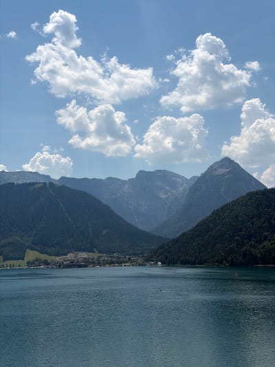 The image depicts a serene lakeside view with a village nestled at the foot of mountains. The foreground is dominated by a large, calm body of blue water, reflecting the bright blue sky and scattered white clouds. The water exhibits subtle ripples, suggesting a gentle breeze. In the middle ground, a shoreline town, identified as Pertisau, Austria, is visible. The town features a collection of buildings with red-tiled roofs, nestled amongst trees and green hills. Beyond the town, a range of majestic, tree-covered mountains rises sharply, their peaks partially obscured by haze. The sky is a vibrant blue, with numerous fluffy white cumulus clouds scattered across it, some casting soft shadows on the landscape below. The overall impression is one of natural beauty and tranquility.