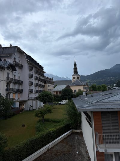 The image shows a view of Saint-Gervais-les-Bains, France, under a cloudy sky. In the foreground, a building with a white facade and multiple balconies with wrought-iron railings is visible. Adjacent to it, another building with a grey pitched roof is partially visible. Further into the scene, a church with a tall, multi-tiered steeple stands prominently. Beyond the church, a residential building with yellow and beige walls and blue shutters is situated. In the background, lush green mountains are partially obscured by clouds, with snow-capped peaks visible in the distance. A well-maintained lawn and trees are present in the middle ground. A white van is parked on a street near the church. The overall impression is one of a serene, mountainous town on an overcast day.