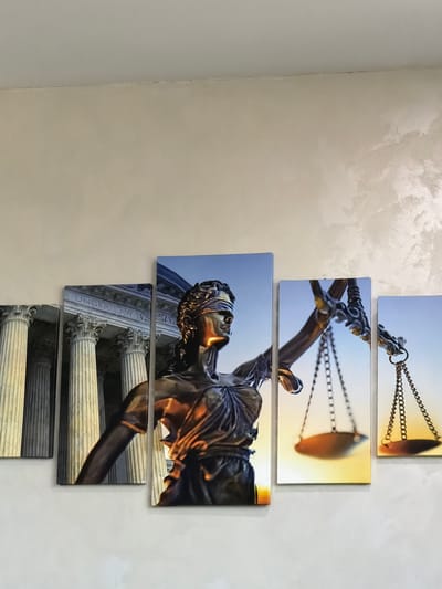A multi-panel canvas print depicting the Statue of Justice is displayed on a light-colored, textured wall. The print features the blindfolded statue holding scales, with images of classical columns and the inscription "E. UNDER LAW" on the left panels. The background of the print transitions from a blue sky at the top to a warm orange and yellow sunset or sunrise at the bottom. The print is artfully arranged with the panels in staggered heights and widths, creating a dynamic visual effect. The wall itself appears to be painted with a decorative plaster technique, adding a subtle shimmer and texture. The lighting suggests an indoor setting, likely a room in a residential or commercial building within the city of Orhei, Moldova.