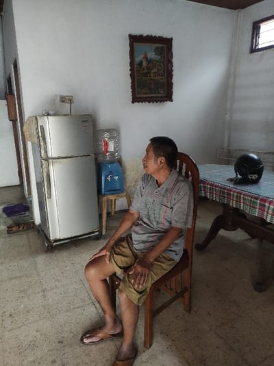 The image shows an adult male, appearing middle-aged or elderly, seated on a wooden chair in an indoor setting. He is dressed in a light grey t-shirt, light brown shorts, and brown flip-flops. His posture suggests he is leaning forward, with both hands resting on his thighs, and he is looking downwards, possibly indicating discomfort, fatigue, or contemplation.
The scene is a modest room, likely a living or dining area in a home in Lamongan, Indonesia. The walls are painted white, and the floor appears to be a light-colored, possibly concrete surface. Natural light illuminates the room, streaming in from a high window on the upper left, suggesting it is daytime.
Various objects are visible around the man. To his right, a wooden dining table with a red, white, and green checkered tablecloth has a black motorcycle helmet resting on it. Behind him and to his left, there is a silver two-door refrigerator. On top of a blue stand or unit adjacent to the refrigerator, a large, inverted clear water bottle is placed in a blue dispenser. A framed landscape painting, depicting houses and trees, hangs on the left wall. No visible text is clearly discernible in the image.