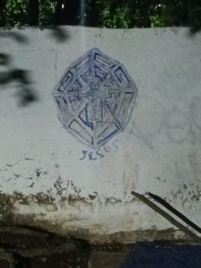 The image depicts a light-colored wall on which graffiti has been drawn. The graffiti consists of a geometric shape, resembling a complex, multi-layered octagon, with a cross at its center. Below the geometric shape, the name "Jesus" is written in a stylized, handwritten font. The wall shows signs of weathering and age, with visible stains and discoloration, especially towards the bottom. In the foreground, partially obscuring the lower right corner of the wall, are two dark, stick-like objects. The background is out of focus, with trees visible at the top of the frame. The location of this image is Tahlequah, United States of America.