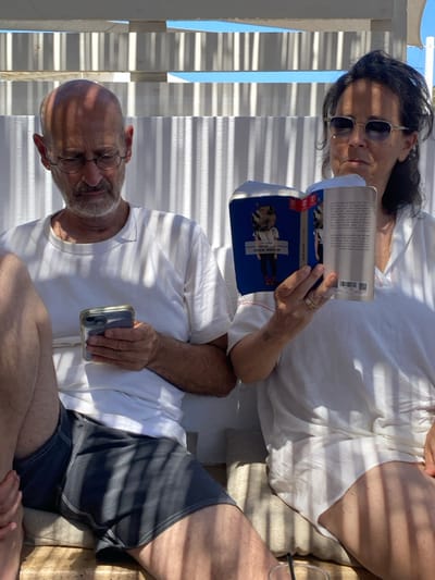 This image depicts two individuals, an adult male and an adult female, seated side-by-side in an outdoor, shaded leisure area in Φάρος, Greece. They are positioned under a white slatted canopy structure that casts distinct parallel shadows across the scene.
The male, situated on the left, is bald with a short beard and wears round-rimmed glasses, a white short-sleeved t-shirt, and dark shorts. He holds a silver-colored smartphone with a clear case in both hands and is looking down at its screen.
The female, on the right, has dark, wavy hair and wears sunglasses and a white, loose-fitting top. She is holding an open book with a prominent blue cover that features an illustration of a figure with a rhinoceros head. The visible text on the book cover and pages is in Hebrew, including the title "ארבעה אבות" (Arba'ah Avot). Her left hand, adorned with a gold-colored ring on the ring finger, also shows a small tattoo of three vertical lines on the forearm. She is looking slightly upward and to the right, not directly at the book.
Both individuals are seated on light-colored cushions positioned on what appears to be a wooden bench. A clear blue sky is visible above the white canopy structure, indicating a bright, sunny day. A portion of a clear glass with a dark straw is visible in the bottom right corner of the frame.
