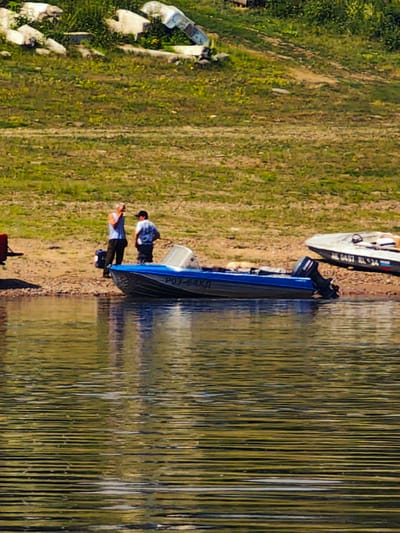 Two men stand on a sandy bank next to a blue motorboat docked at the water's edge. The man on the left wears a light blue sleeveless shirt and dark pants, holding a dark object to his ear, possibly a phone. The man on the right wears a dark shirt with a white pattern and dark pants, and has a baseball cap on his head. The blue motorboat has the registration number "P03-64XH" visible on its side. Further to the right, a second motorboat, white with a dark stripe and registration "NE 0497 RL$24", is also beached. The background consists of a grassy slope with scattered white debris and vegetation. The water in the foreground is rippled and reflects the surrounding environment. The scene appears to be outdoors, likely near a body of water, given the presence of boats and the shoreline. The location of Погодаево, Russia, is not discernible from the visual elements within the image itself.