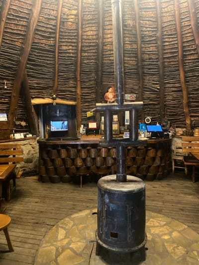 The image depicts the interior of a circular or conical building, identified as being in Dulovine, Montenegro. The structure features a high ceiling and walls composed of numerous horizontal layers of dark wooden sticks or branches, supported by vertical dark wooden beams. Centrally positioned in the foreground is a dark, cylindrical metal wood-burning stove, from which a multi-sectioned, dark metal stovepipe extends vertically towards the ceiling. This stove rests on a semi-circular patch of irregular stone tiling embedded in the wooden plank floor.
Behind the stove, a semi-circular bar or counter is visible, its front facade constructed from stacked, dark-stained wooden cylindrical sections. On the bar surface, from left to right, are various items including what appears to be a red-framed sign, a vintage radio, and a modern point-of-sale system with an illuminated blue screen. A framed portrait of a woman in traditional attire hangs on the wall above the bar, flanked by a small "No Smoking" sign to its immediate left.
To the left of the bar, a dark wooden archway with a mounted horseshoe leads into another room, where a television screen displaying an image is visible. On the far left and right sides of the image, sections of wooden tables and benches are visible, suggesting a seating area. The overall lighting is dim. No individuals are present in the scene.