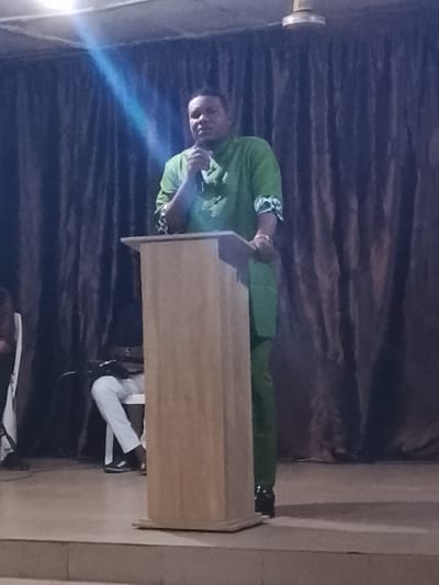 A man in a green suit stands behind a light brown podium on a stage with a shiny surface. He holds a microphone, speaking to an unseen audience. Behind him are dark brown curtains. To the left, a person in white pants and black shoes sits on a chair. A bright light shines from above, creating a glare on the image.