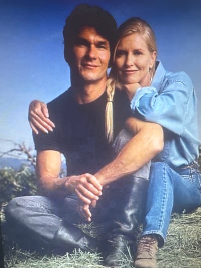 The image shows a medium shot of a man and a woman sitting outdoors. The man, in the foreground, is wearing a black t-shirt and dark jeans. He is looking at the camera with a slight smile. The woman is sitting to his right, leaning her head on his shoulder. She is wearing a light blue denim shirt and blue jeans. Her hair is blonde and styled in a braid. Both are seated on a grassy area with a blue sky in the background. There are no visible elements that indicate a location in Protasovo, Russia.